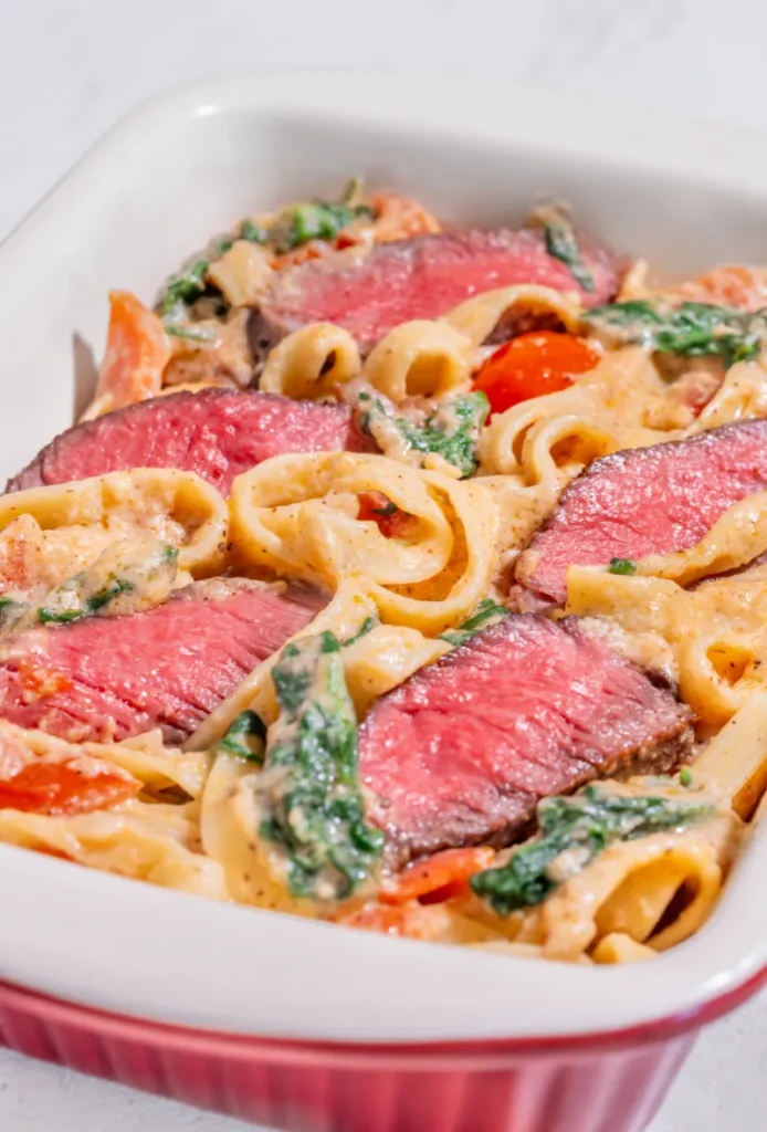 Vertical image of creamy steak fettuccine served in a red oval dish, featuring thick slices of medium-rare steak, fettuccine pasta, spinach, and cherry tomatoes in a rich, creamy sauce.