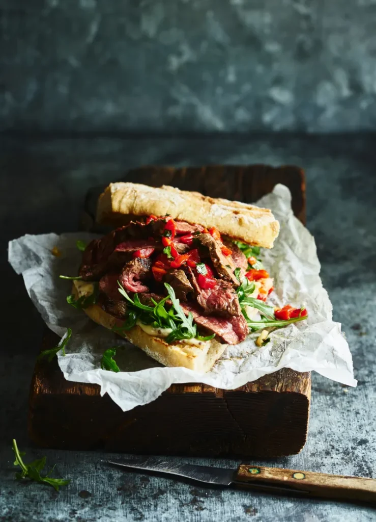 Gourmet steak sandwich on a rustic wooden board, featuring thinly sliced medium-rare steak, fresh arugula, and red chili peppers on a crusty baguette.