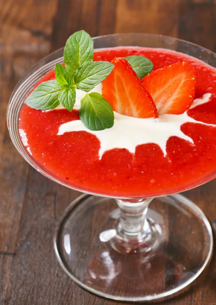 Close-up view of a dessert glass filled with strawberry puree, topped with a swirl of cream, fresh strawberry slices, and mint leaves on a wooden table.