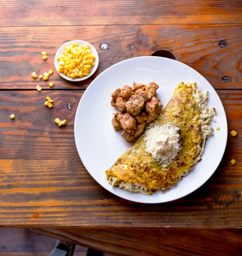 A plate featuring a grilled cachapa topped with cheese and sour cream next to crispy fried meat chunks, with a small bowl of corn kernels beside it.