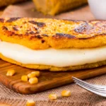 A grilled cachapa filled with melted cheese, served on a wooden board with scattered corn kernels and a fork beside it.