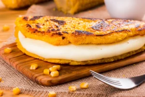 A grilled cachapa filled with melted cheese, served on a wooden board with scattered corn kernels and a fork beside it.