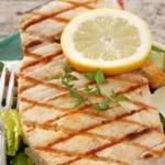 Close-up of a grilled swordfish steak garnished with a lemon slice and fresh herbs, served on a bed of Caesar salad with croutons and shaved Parmesan cheese.