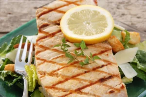 Close-up of a grilled swordfish steak garnished with a lemon slice and fresh herbs, served on a bed of Caesar salad with croutons and shaved Parmesan cheese.