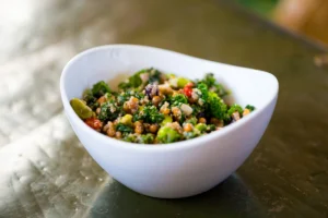 Close-up of a kale and quinoa salad in a white, asymmetric bowl. The salad includes broccoli, beans, and diced vegetables, offering a variety of colors like green, red, and yellow, placed on a scratched metal surface.