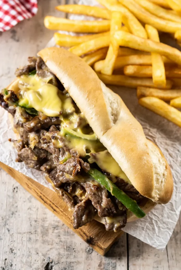 A Philly cheesesteak sandwich with melted cheese and green bell peppers on a hoagie roll, served with a side of golden fries on a rustic wooden board.