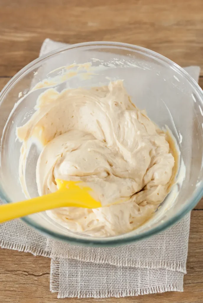 A glass bowl filled with freshly made Russian buttercream, with a yellow silicone spatula folding the mixture on a wooden table.