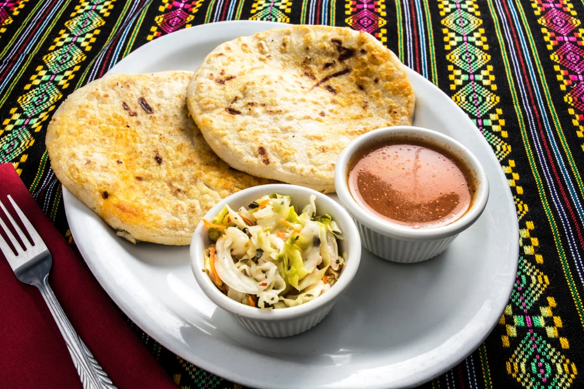 Two pupusas with a side of curtido and salsa on a white plate.