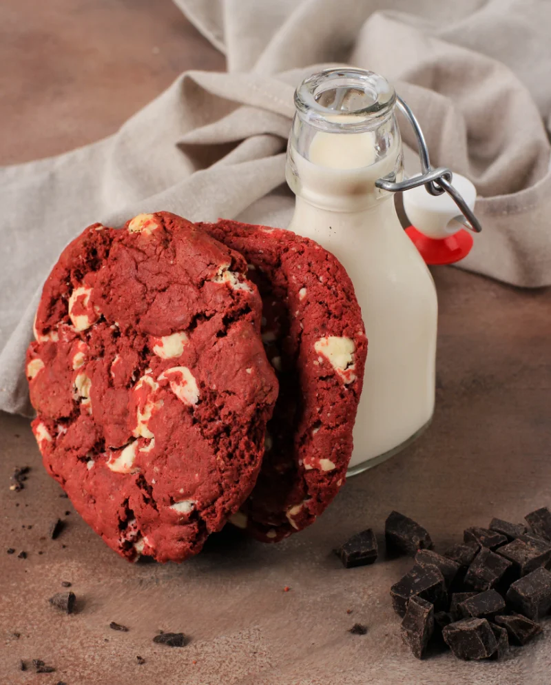 Stack of red velvet cookies with white chocolate chips next to a small glass milk bottle, with dark chocolate pieces and a grey fabric in the background.