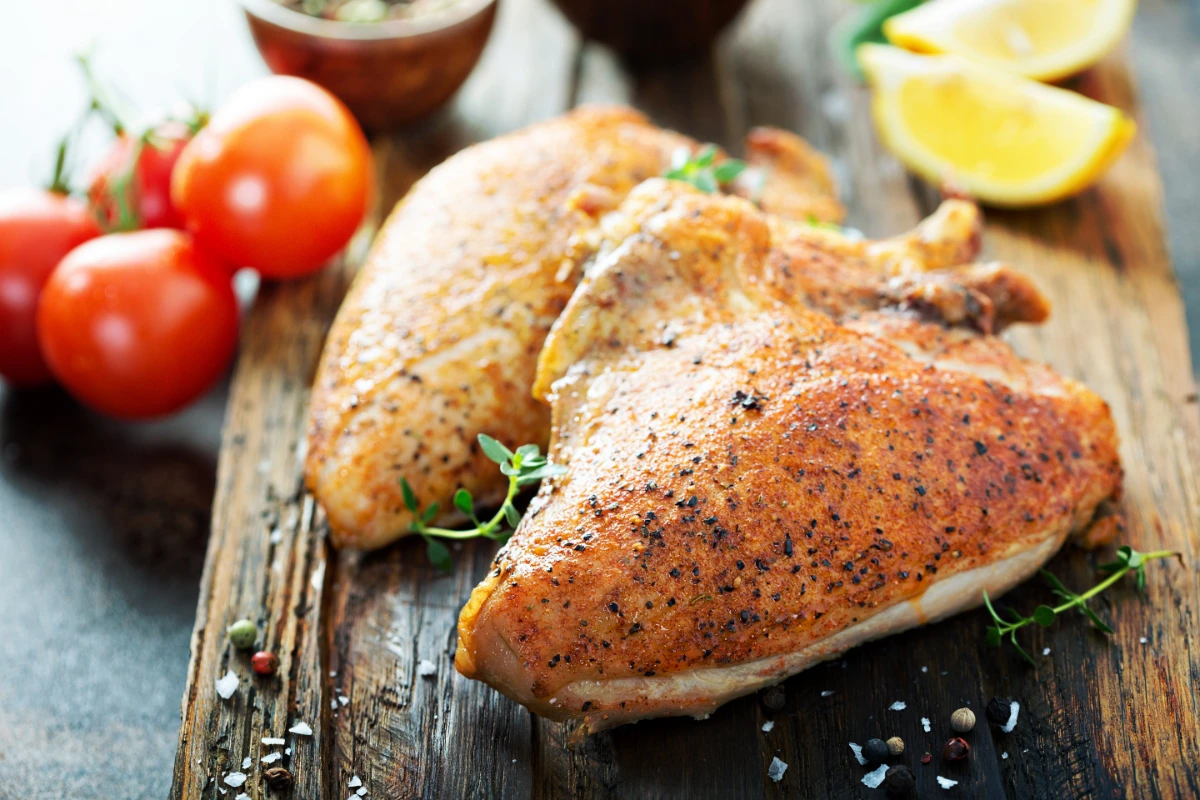 Two seasoned and smoked chicken breasts on a wooden cutting board, garnished with fresh thyme. Accompanied by whole cherry tomatoes, scattered peppercorns, sea salt, and lemon wedges in the background.