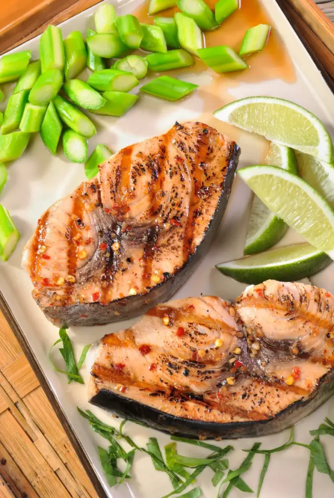 Two spiced swordfish steaks with grill marks served on a rectangular plate, accompanied by chopped snap peas and lime wedges.