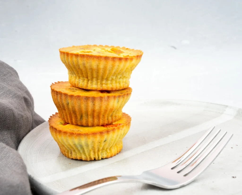 Three golden egg bites stacked on a white ceramic plate next to a fork, with a light gray napkin partially visible.