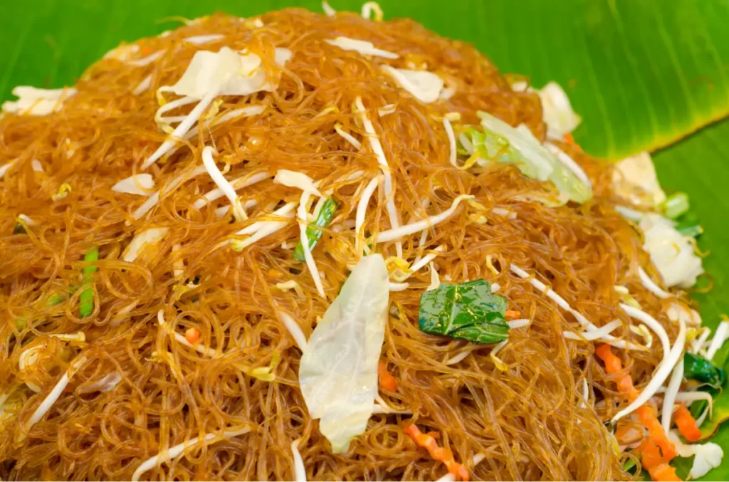 A plate of stir-fried vermicelli with bean sprouts, cabbage, and green vegetables on a green background.