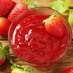 Close-up of a bowl of strawberry puree surrounded by whole strawberries on a wooden surface.