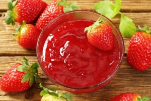 Close-up of a bowl of strawberry puree surrounded by whole strawberries on a wooden surface.