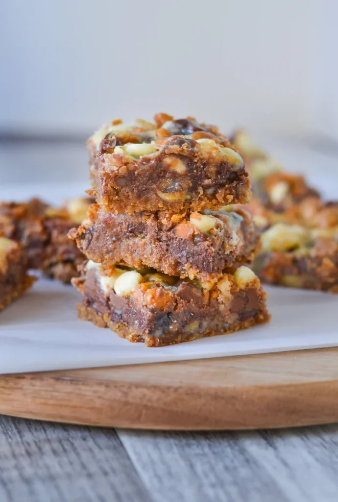 A stack of three 7 layer bars with visible layers of nuts, chocolate, and coconut on a white plate set on a wooden cutting board.