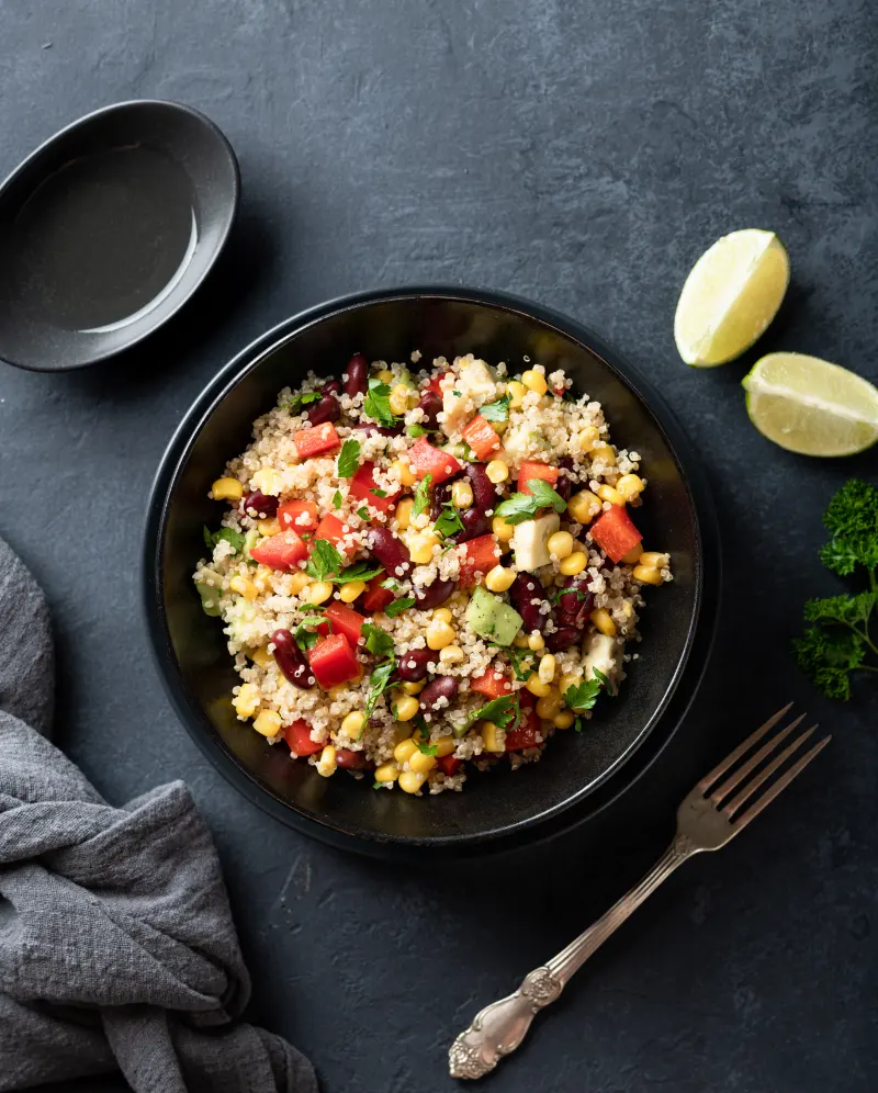 Zesty quinoa salad served in a dark bowl on a slate gray background. The salad is a colorful mix of red kidney beans, diced tomatoes, sweet corn, and fresh herbs, garnished with lime wedges. A small black bowl and an antique fork rest beside the main dish, enhancing the rustic aesthetic.