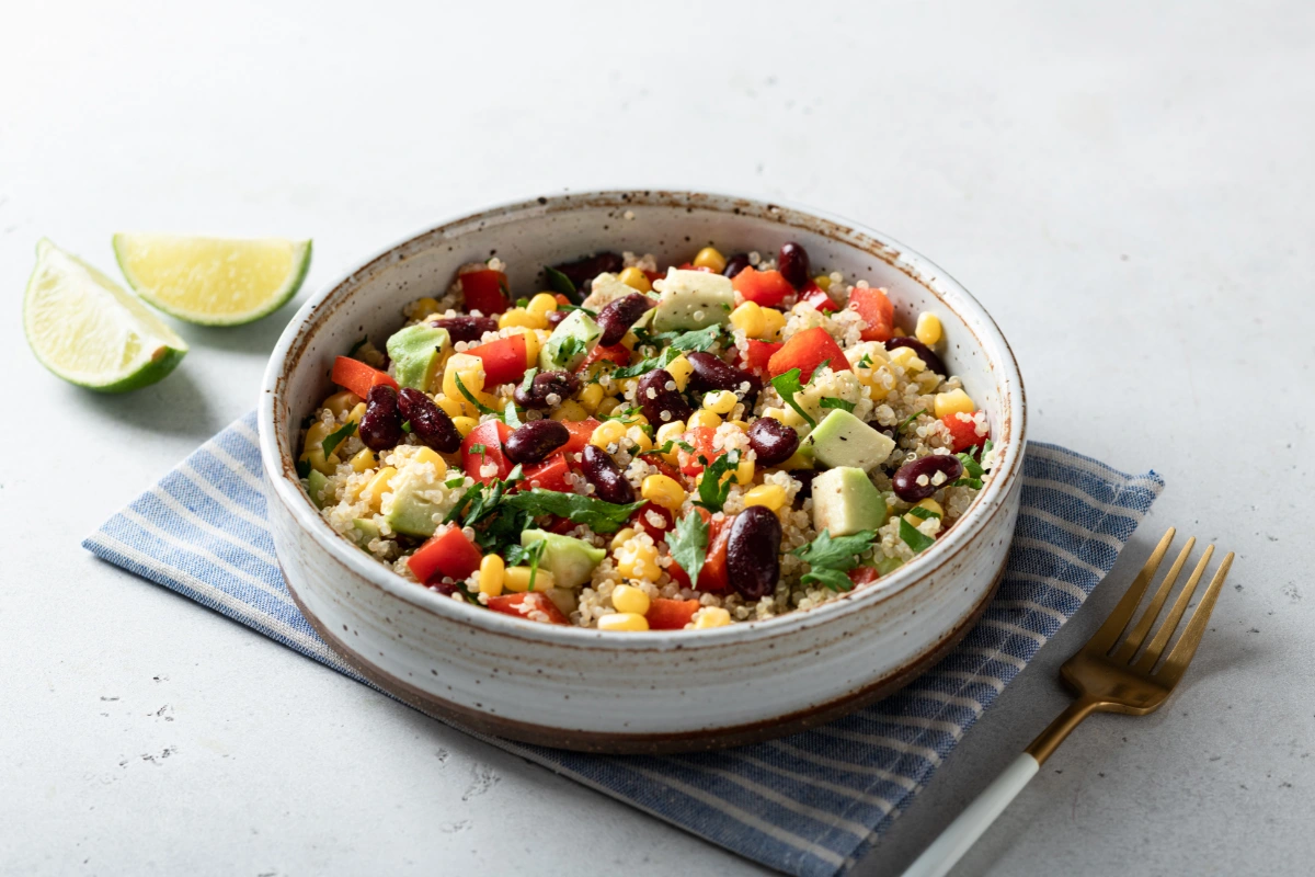 A zesty quinoa salad in a speckled ceramic bowl, featuring diced avocado, red kidney beans, sweet corn, red bell peppers, and chopped parsley. Two lime wedges are placed on the left side of the bowl, which rests on a blue-striped cloth with a golden fork on a gray surface.