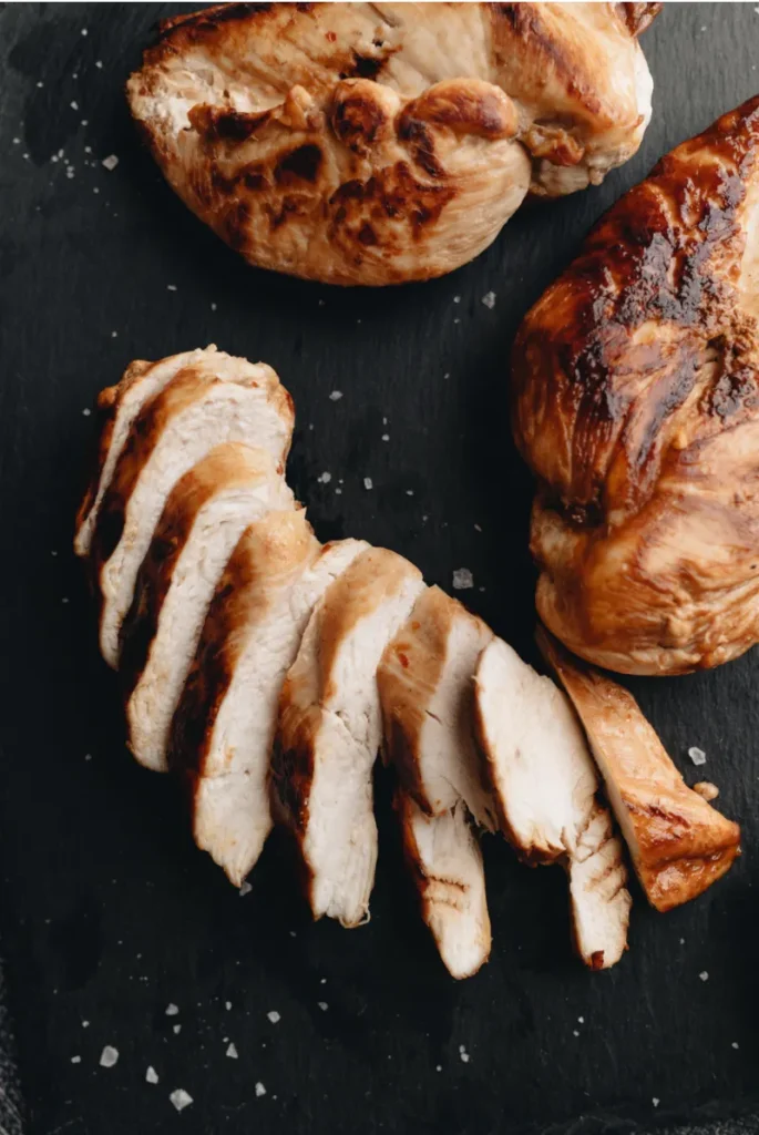 Close-up view of smoked chicken breasts, one sliced into pieces, on a dark slate surface. Visible grill marks on the golden-brown skin and scattered sea salt crystals enhance the texture.