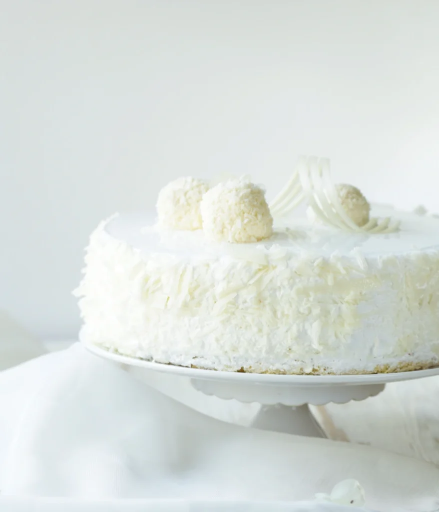 Whole coconut cake topped with coconut balls and white chocolate curls on a cake stand.